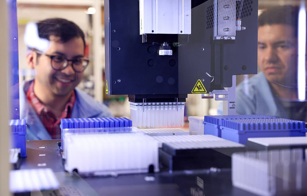 Two scientists watch a machine fill a tray of vials